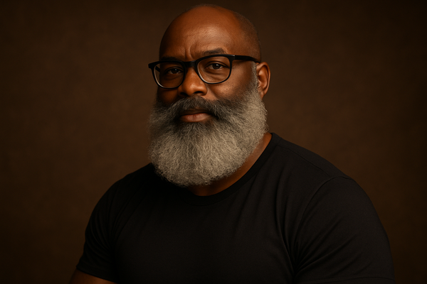 older black man with salt and pepper beard, the beard is BIG fluffy well moisturized and head-turning. the man wears a black t-shirt, his muscles featured prominently, his black glasses frame his face well.